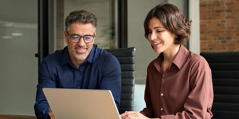 A man and a woman smiling and working together on a laptop at a wooden table in a modern office setting.