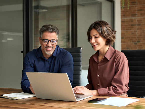 A man and a woman smiling and working together on a laptop at a wooden table in a modern office setting.