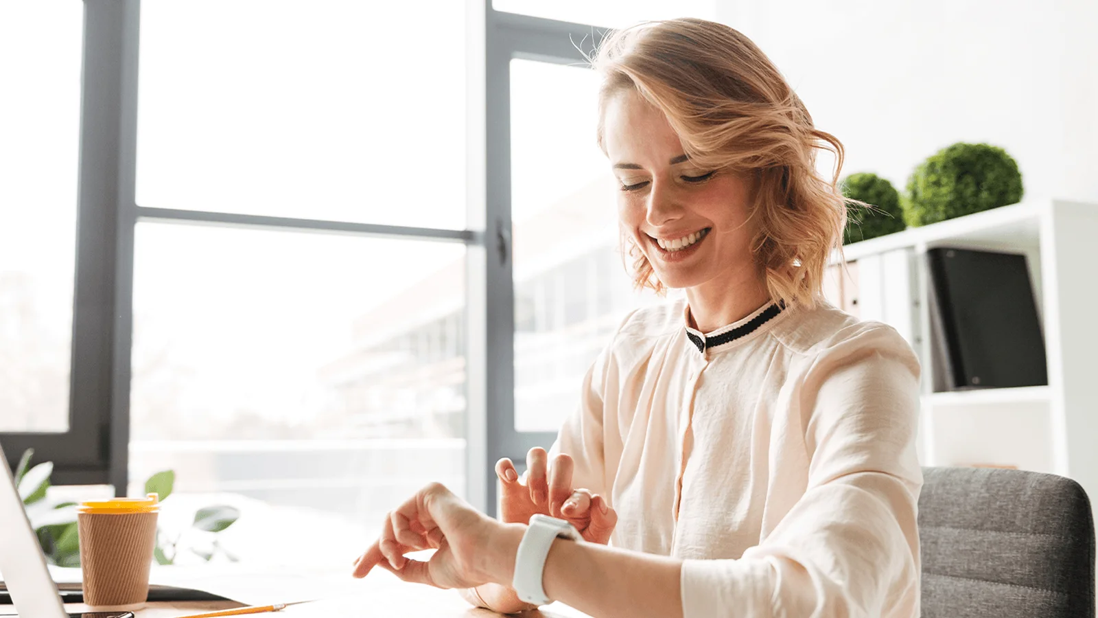 A person sitting at a desk, smiling at a smartwatch. A laptop, glasses, and a coffee cup are on the desk in a bright office space.