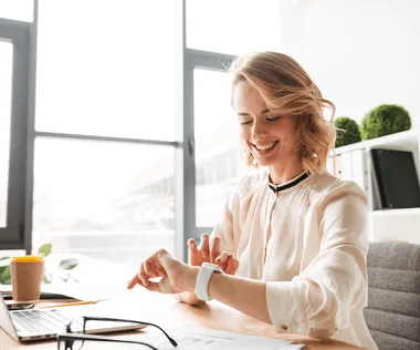 A person sitting at a desk, smiling at a smartwatch. A laptop, glasses, and a coffee cup are on the desk in a bright office space.
