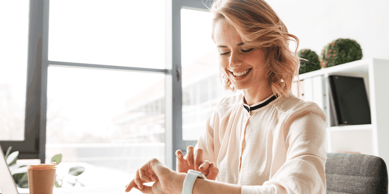 A person sitting at a desk, smiling at a smartwatch. A laptop, glasses, and a coffee cup are on the desk in a bright office space.