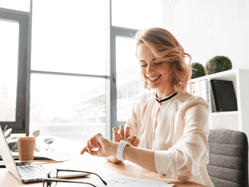 A person sitting at a desk, smiling at a smartwatch. A laptop, glasses, and a coffee cup are on the desk in a bright office space.