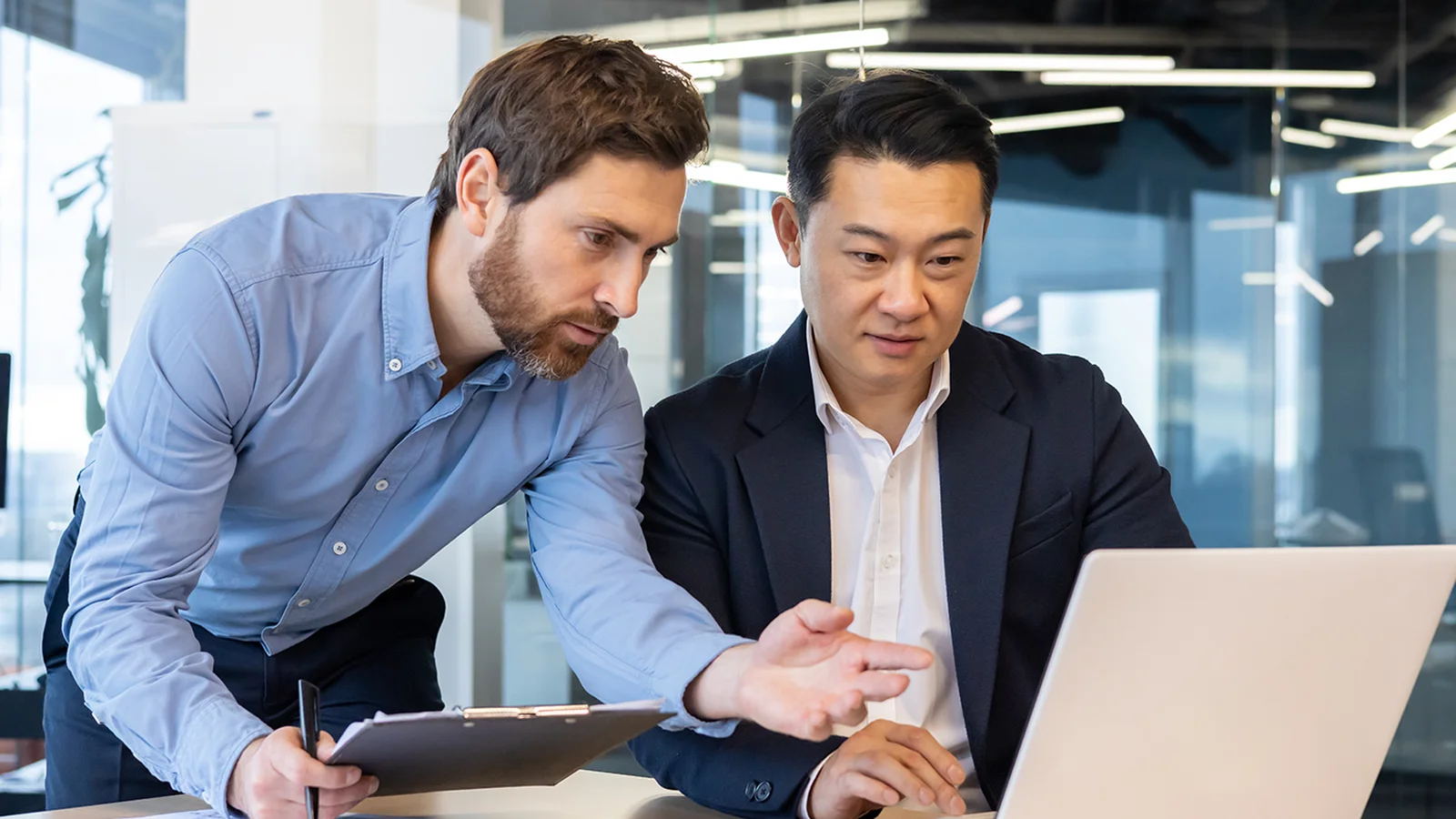 Two men in an office, one pointing at a laptop screen, and the other focused, discussing work.