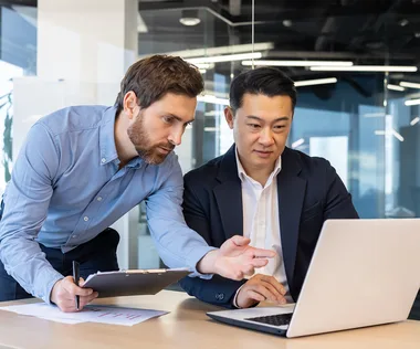 Two men in an office, one pointing at a laptop screen, and the other focused, discussing work.