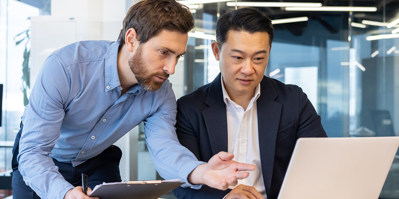 Two men in an office, one pointing at a laptop screen, and the other focused, discussing work.