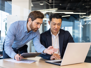 Two men in an office, one pointing at a laptop screen, and the other focused, discussing work.