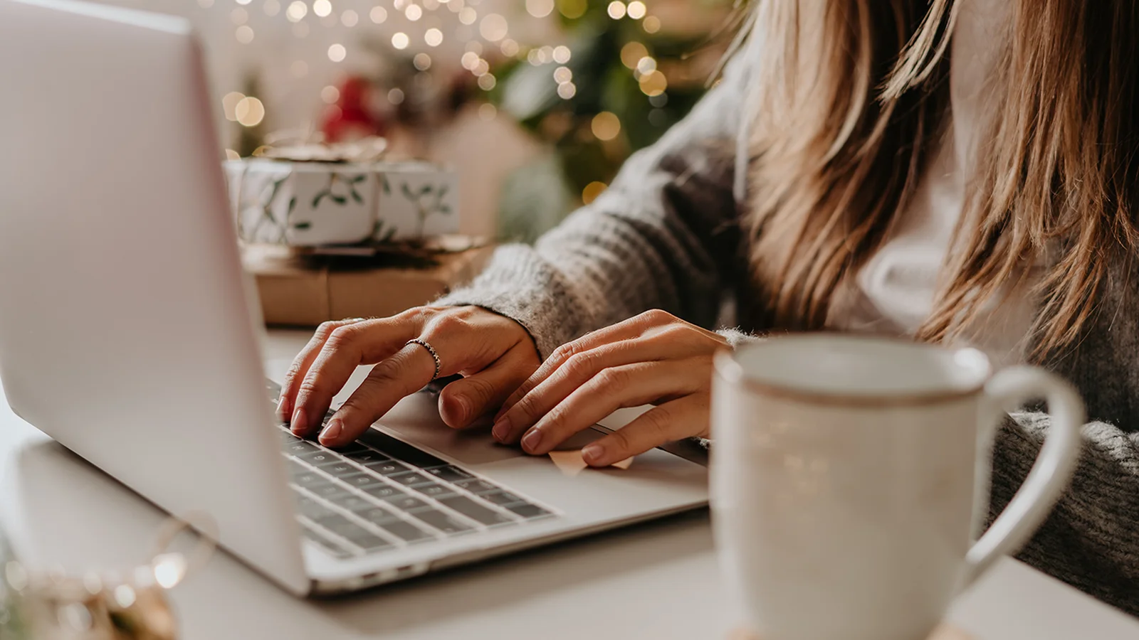 A woman's hands typing on a laptop in a cozy winter holiday setting with a coffee mug next to her.