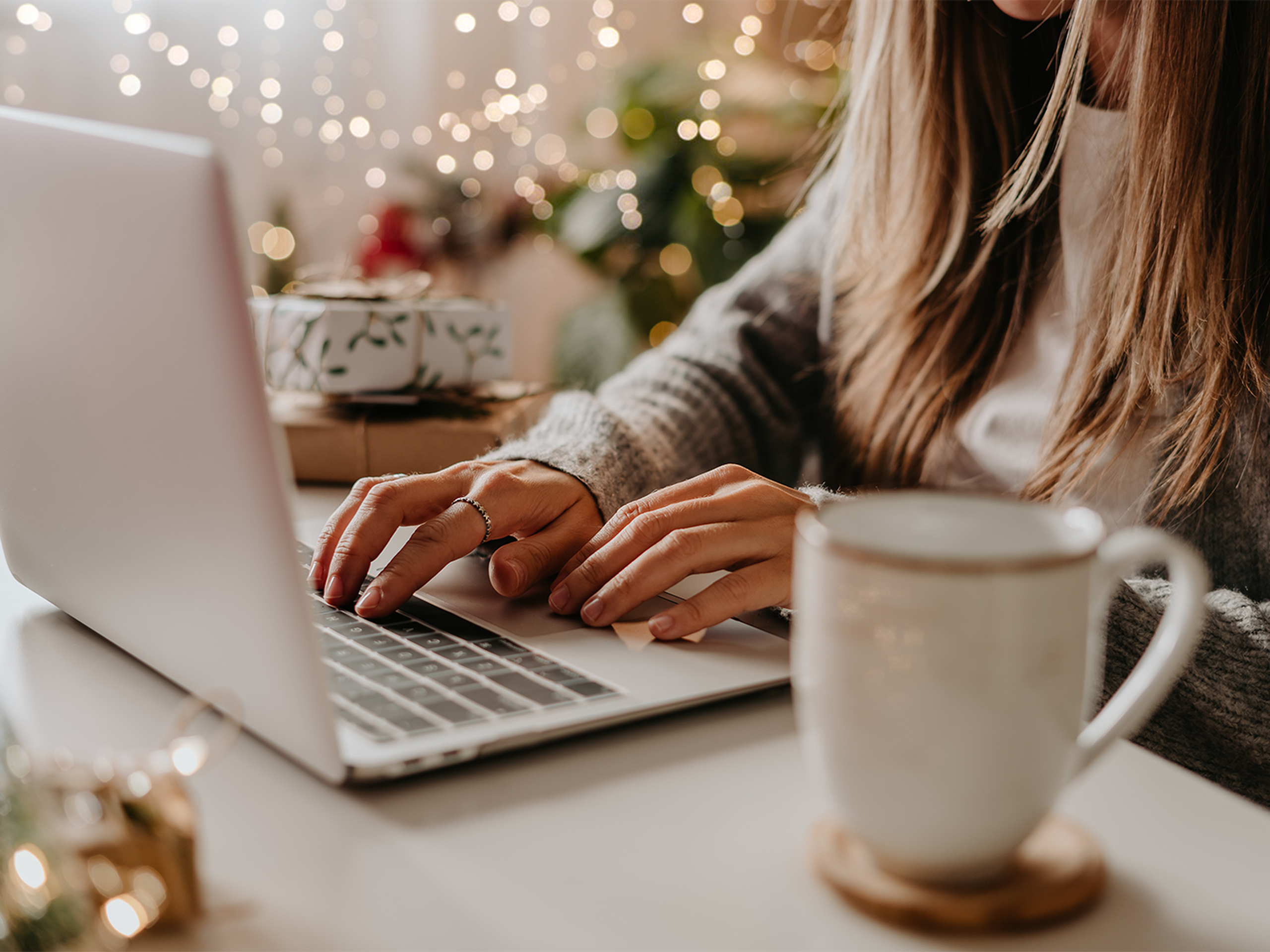 A woman's hands typing on a laptop in a cozy winter holiday setting with a coffee mug next to her.
