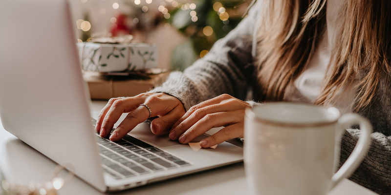A woman's hands typing on a laptop in a cozy winter holiday setting with a coffee mug next to her.