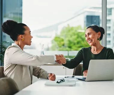 Two women shaking hands in an office setting, seated at a desk with a laptop. Large windows display a blurred cityscape outside.