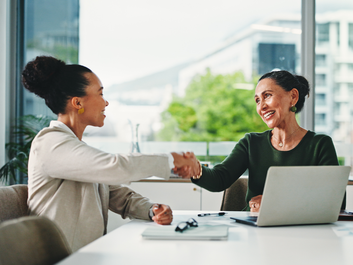 Two women shaking hands in an office setting, seated at a desk with a laptop. Large windows display a blurred cityscape outside.