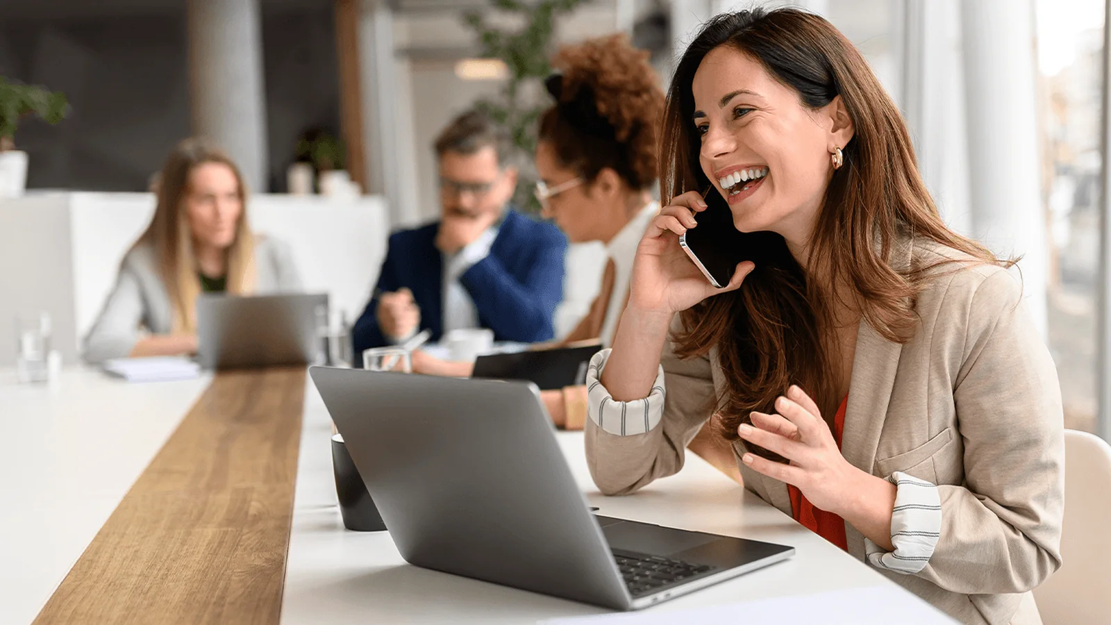 A woman in a beige blazer talks on the phone and smiles while working on a laptop at a modern office, with colleagues in the background.