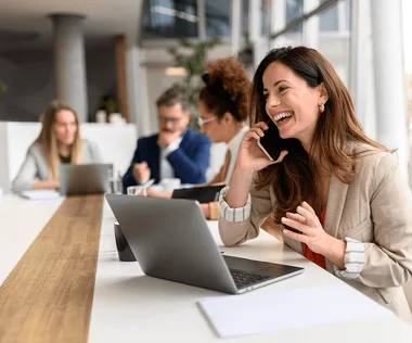 A woman in a beige blazer talks on the phone and smiles while working on a laptop at a modern office, with colleagues in the background.