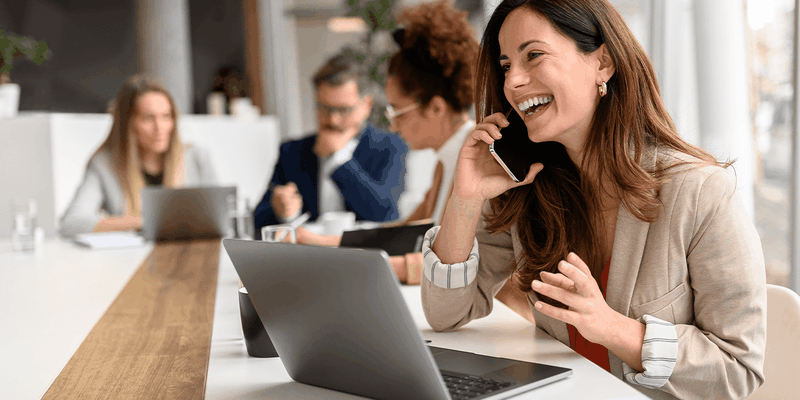 A woman in a beige blazer talks on the phone and smiles while working on a laptop at a modern office, with colleagues in the background.