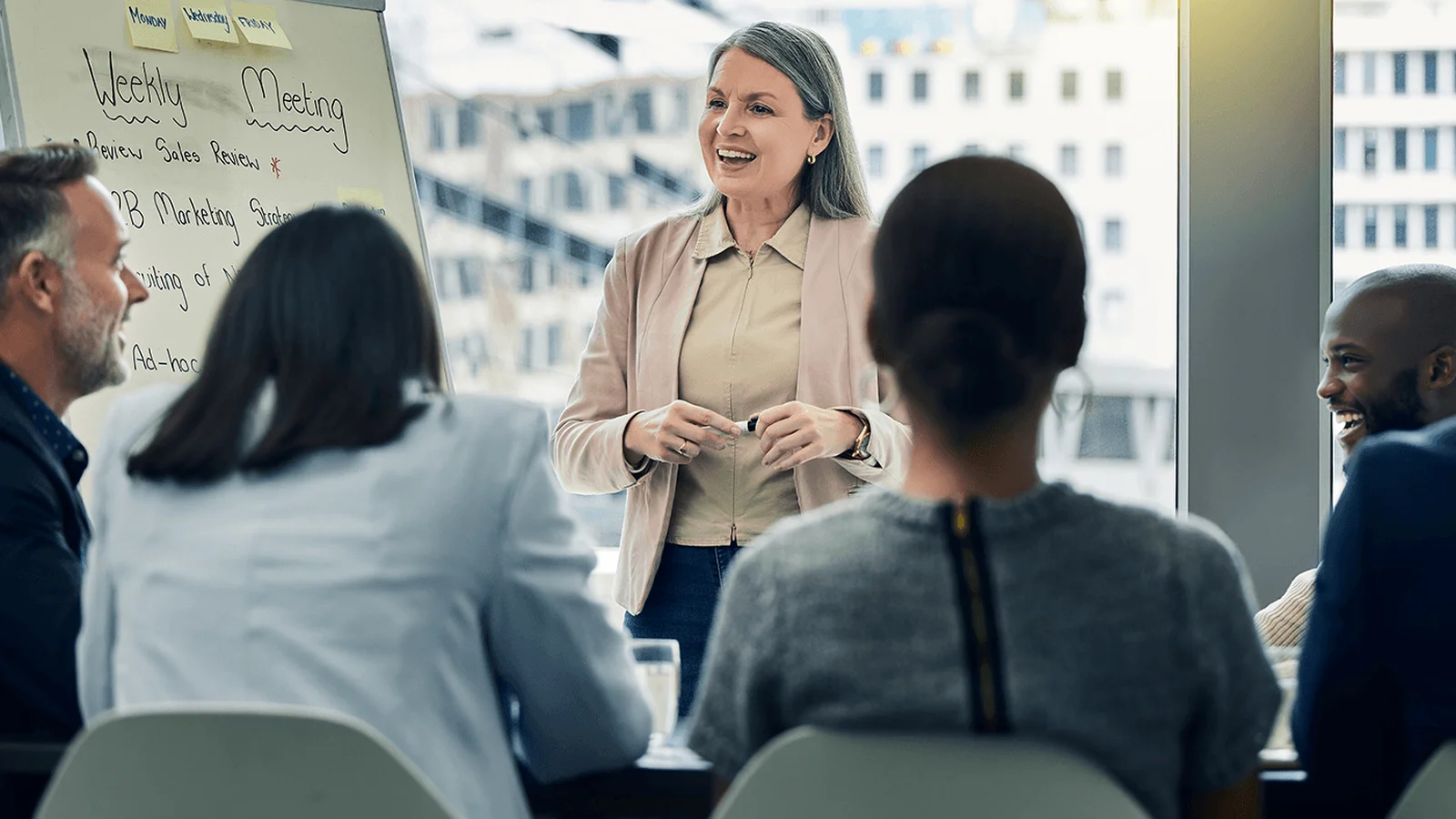 A woman stands and speaks to a group of people seated around a table in a bright office setting, with a whiteboard labeled "Weekly Meeting."