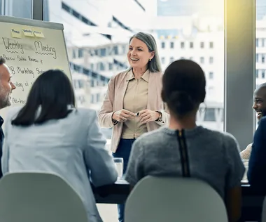 A woman stands and speaks to a group of people seated around a table in a bright office setting, with a whiteboard labeled "Weekly Meeting."