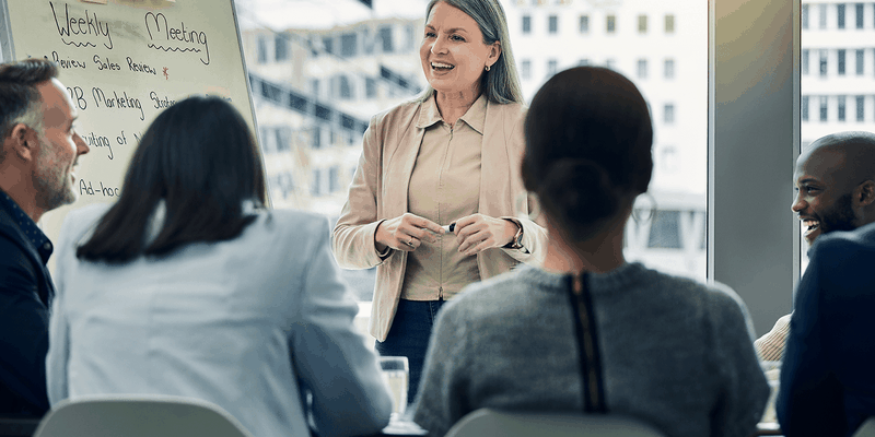 A woman stands and speaks to a group of people seated around a table in a bright office setting, with a whiteboard labeled "Weekly Meeting."