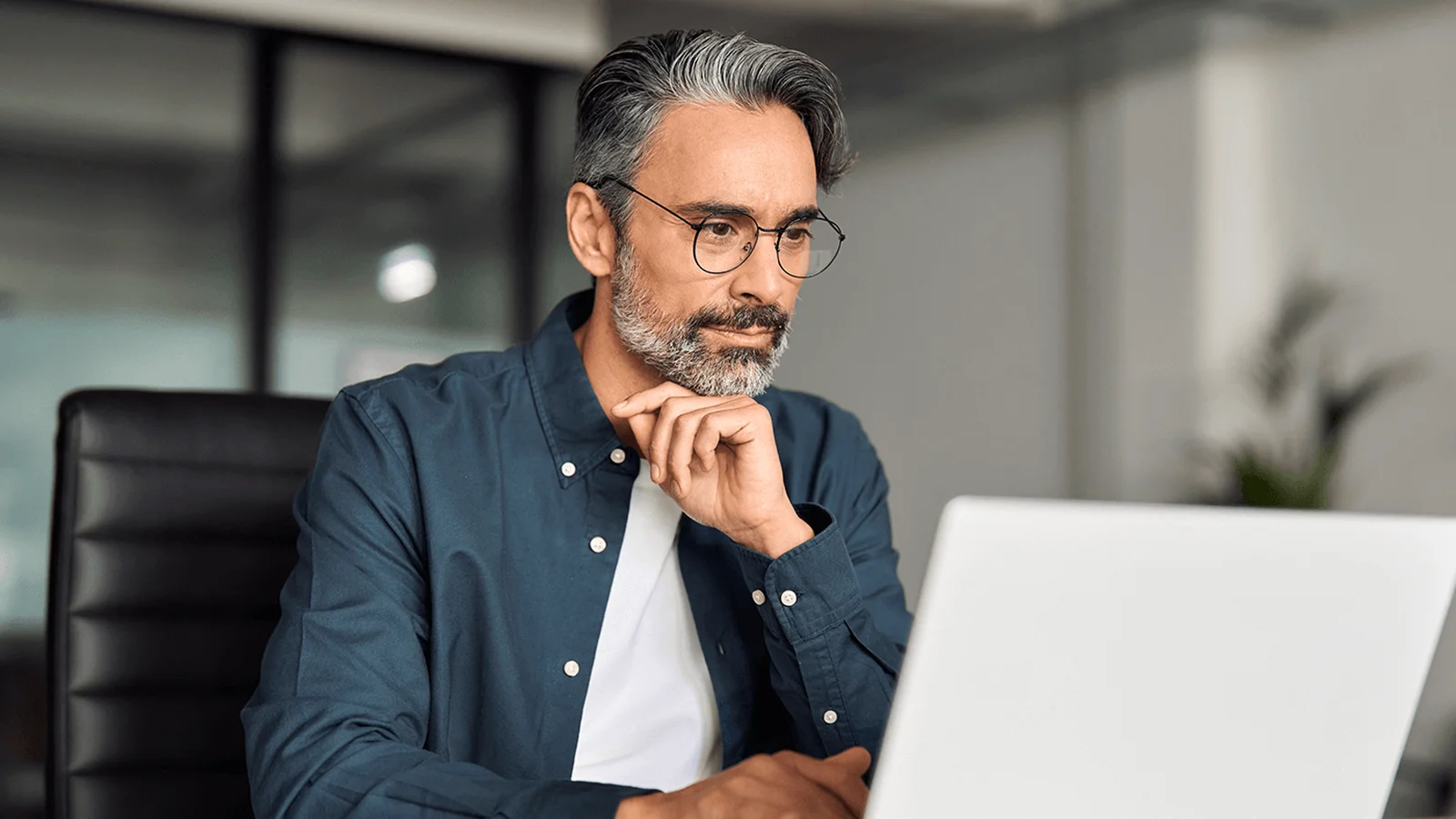 Thoughtful middle-aged man with glasses and gray beard working on a laptop at an office desk.