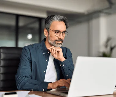 Thoughtful middle-aged man with glasses and gray beard working on a laptop at an office desk.