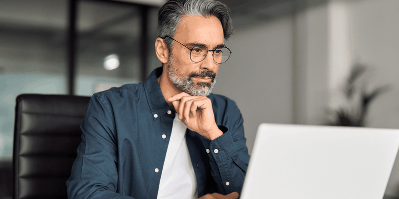 Thoughtful middle-aged man with glasses and gray beard working on a laptop at an office desk.