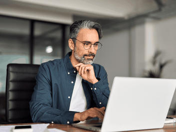 Thoughtful middle-aged man with glasses and gray beard working on a laptop at an office desk.