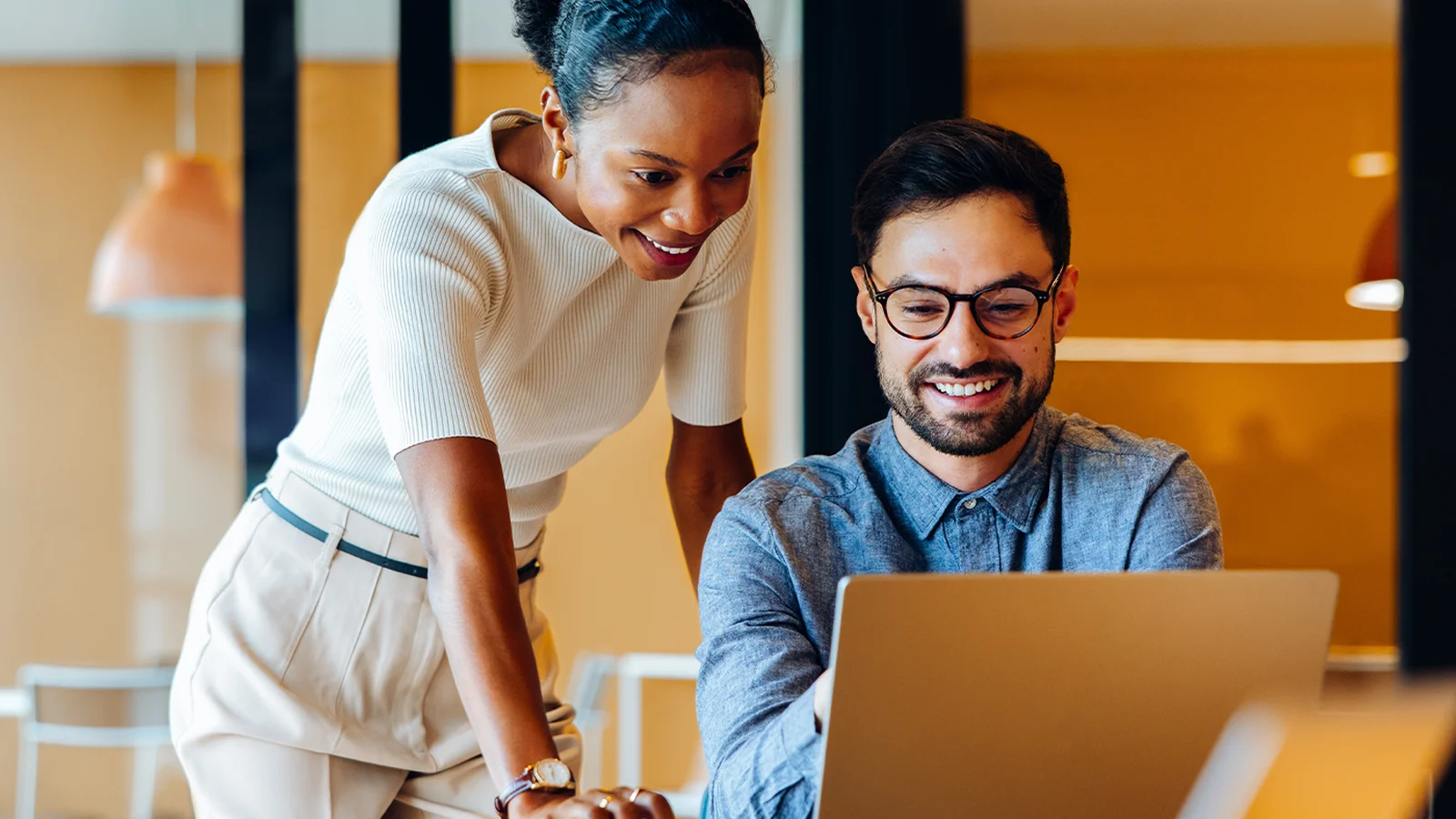 Two colleagues smiling and collaborating at a laptop in a bright office with warm lighting.