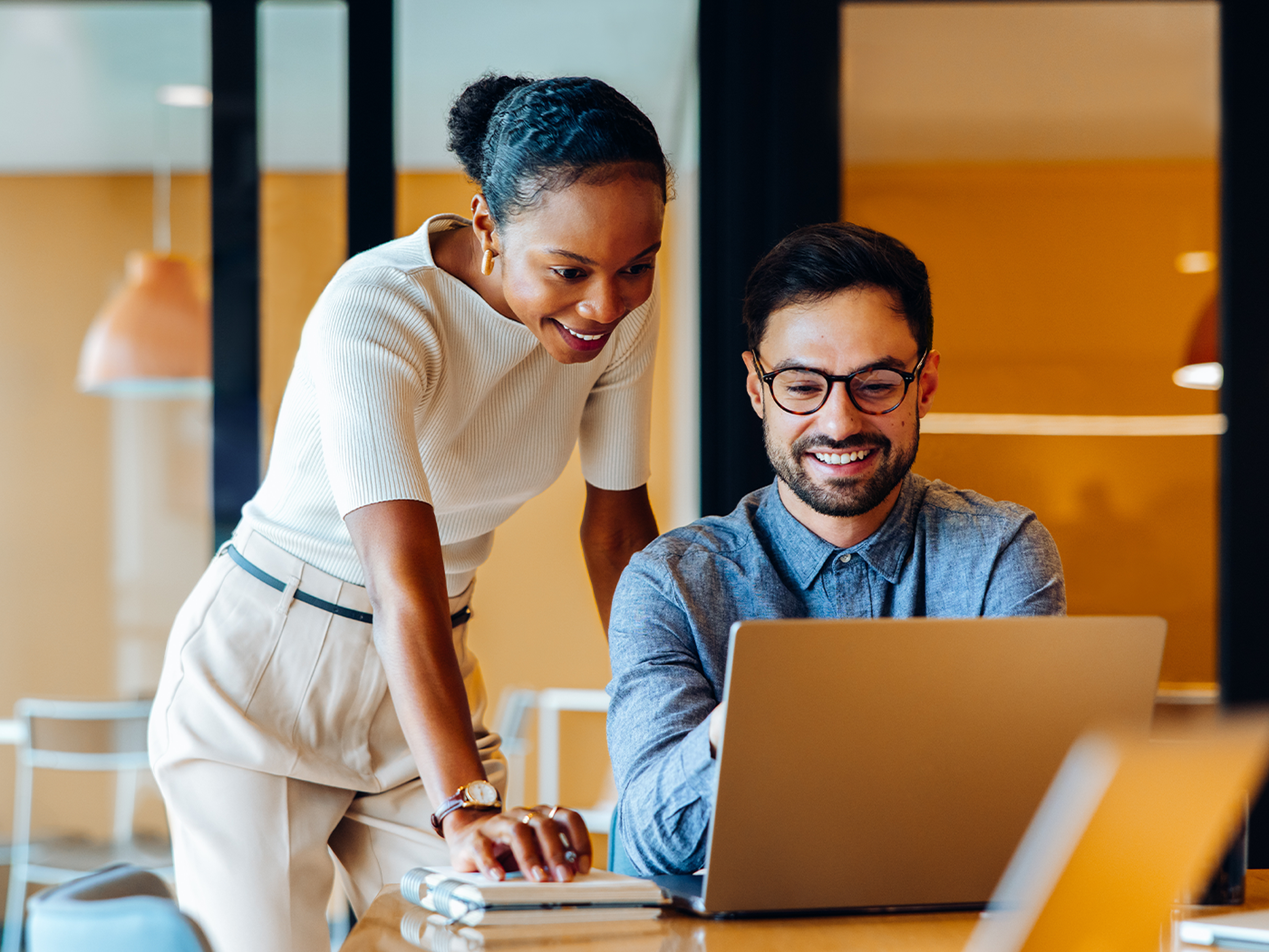 Two colleagues smiling and collaborating at a laptop in a bright office with warm lighting.