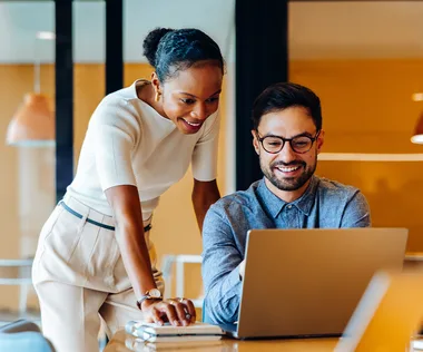Two colleagues smiling and collaborating at a laptop in a bright office with warm lighting.