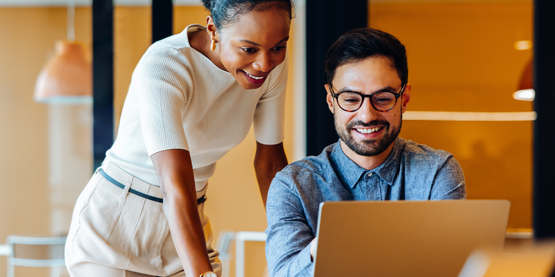 Two colleagues smiling and collaborating at a laptop in a bright office with warm lighting.