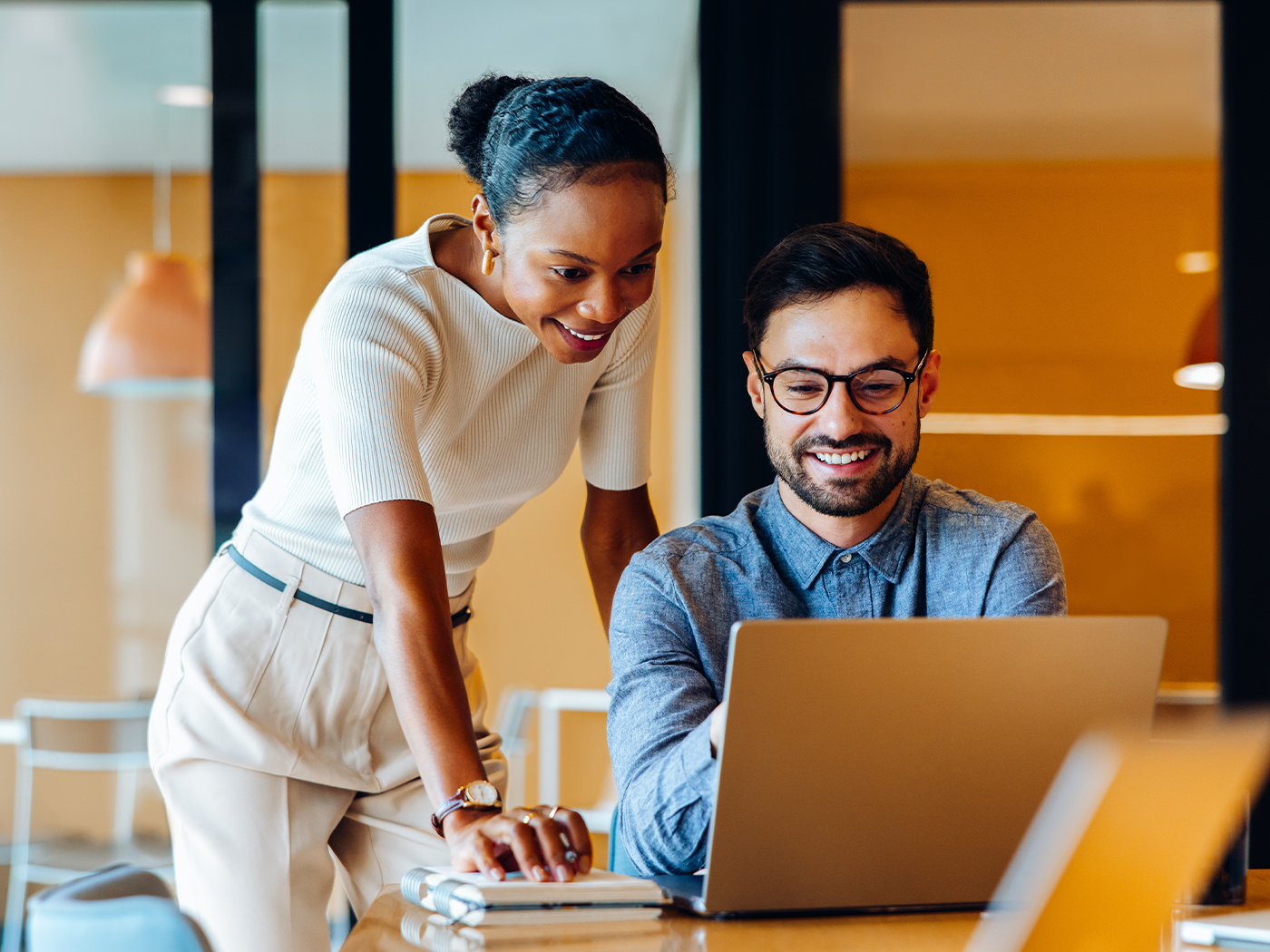Two colleagues smiling and collaborating at a laptop in a bright office with warm lighting.