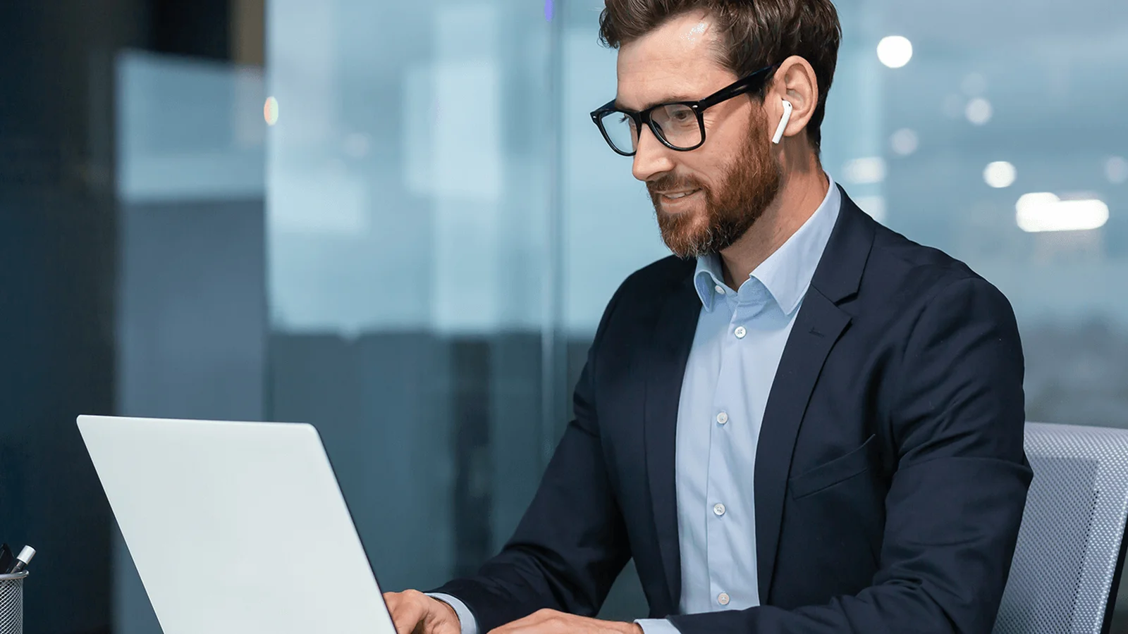 Man in a suit and glasses using a laptop at a desk in a modern office. He is wearing wireless earbuds and smiling slightly.