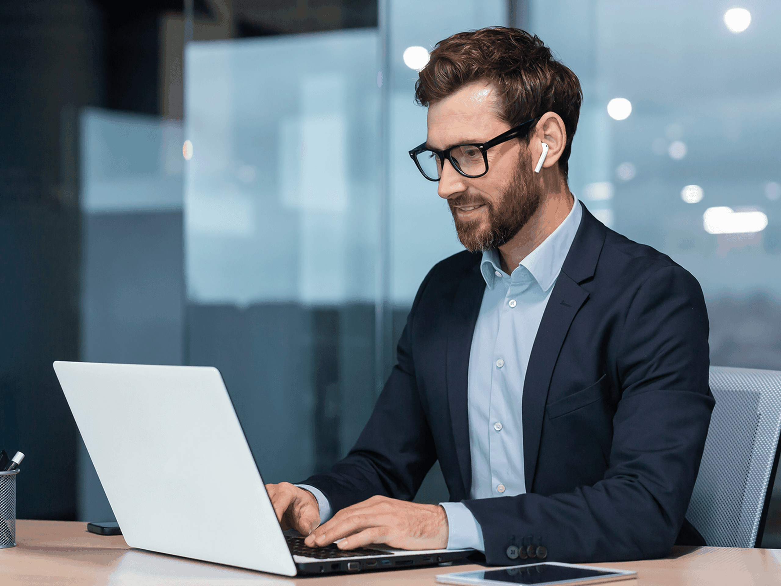 Man in a suit and glasses using a laptop at a desk in a modern office. He is wearing wireless earbuds and smiling slightly.