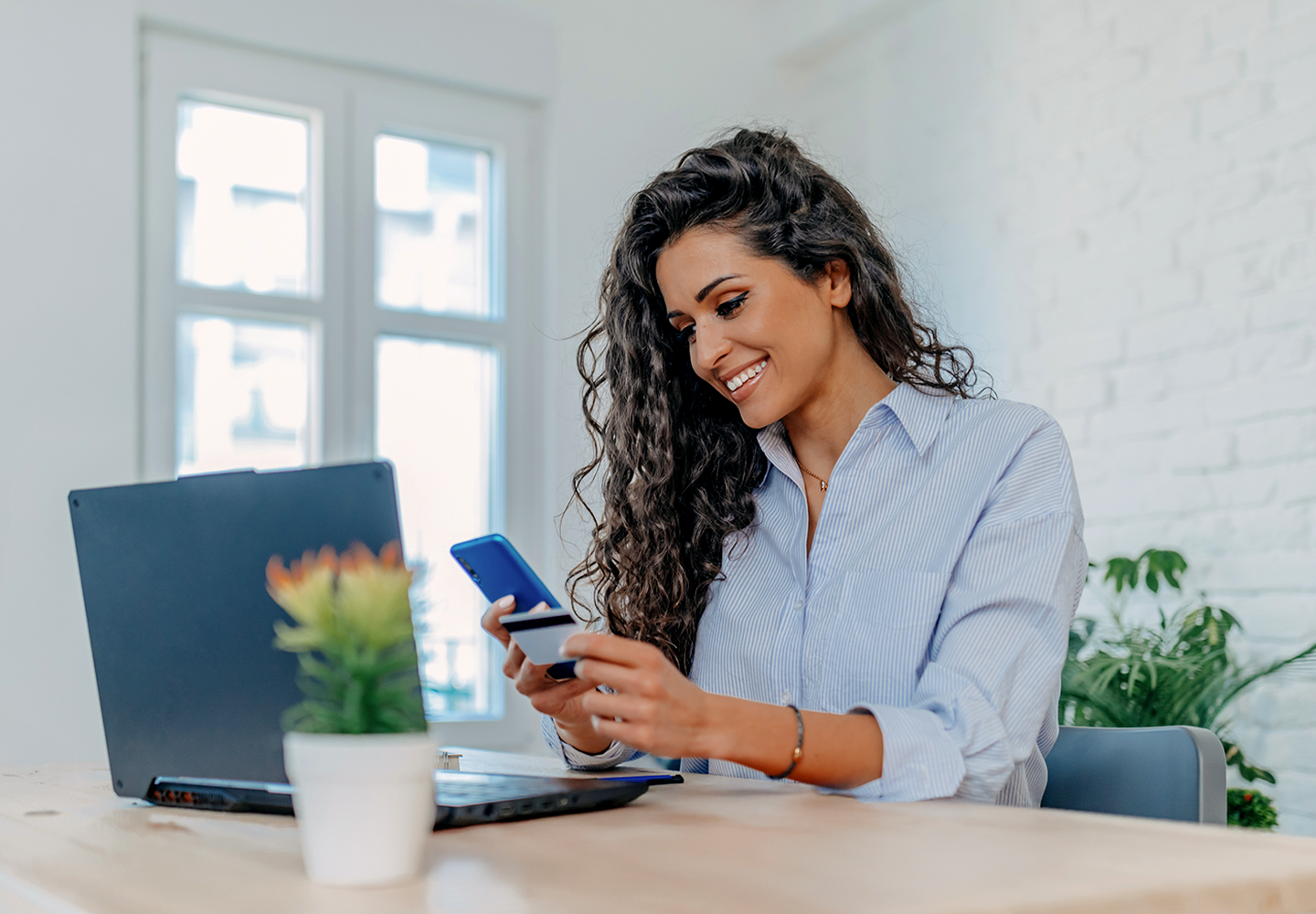 Smiling woman at desk holding a smartphone and credit card, making an online payment beside a laptop.