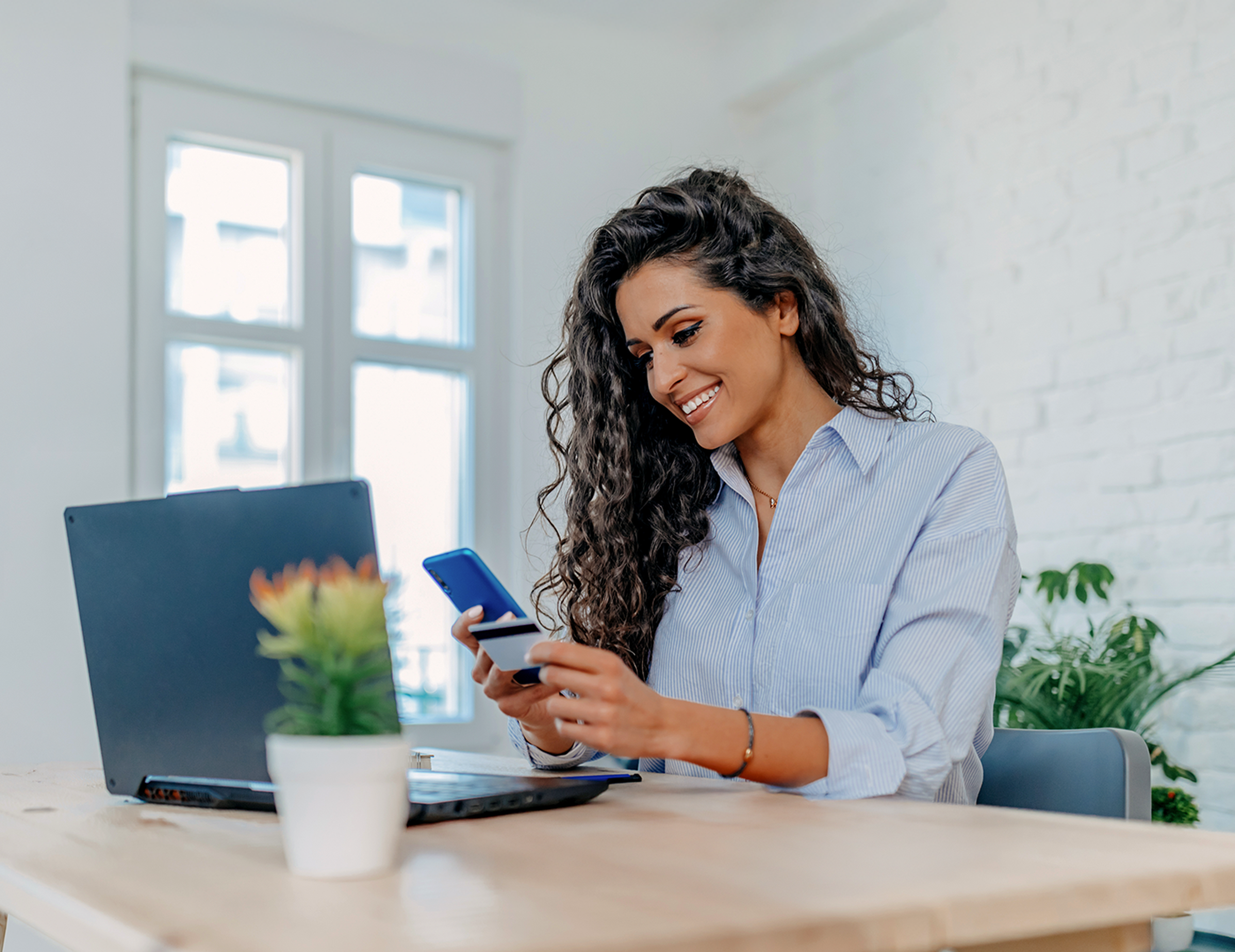 Smiling woman at desk holding a smartphone and credit card, making an online payment beside a laptop.
