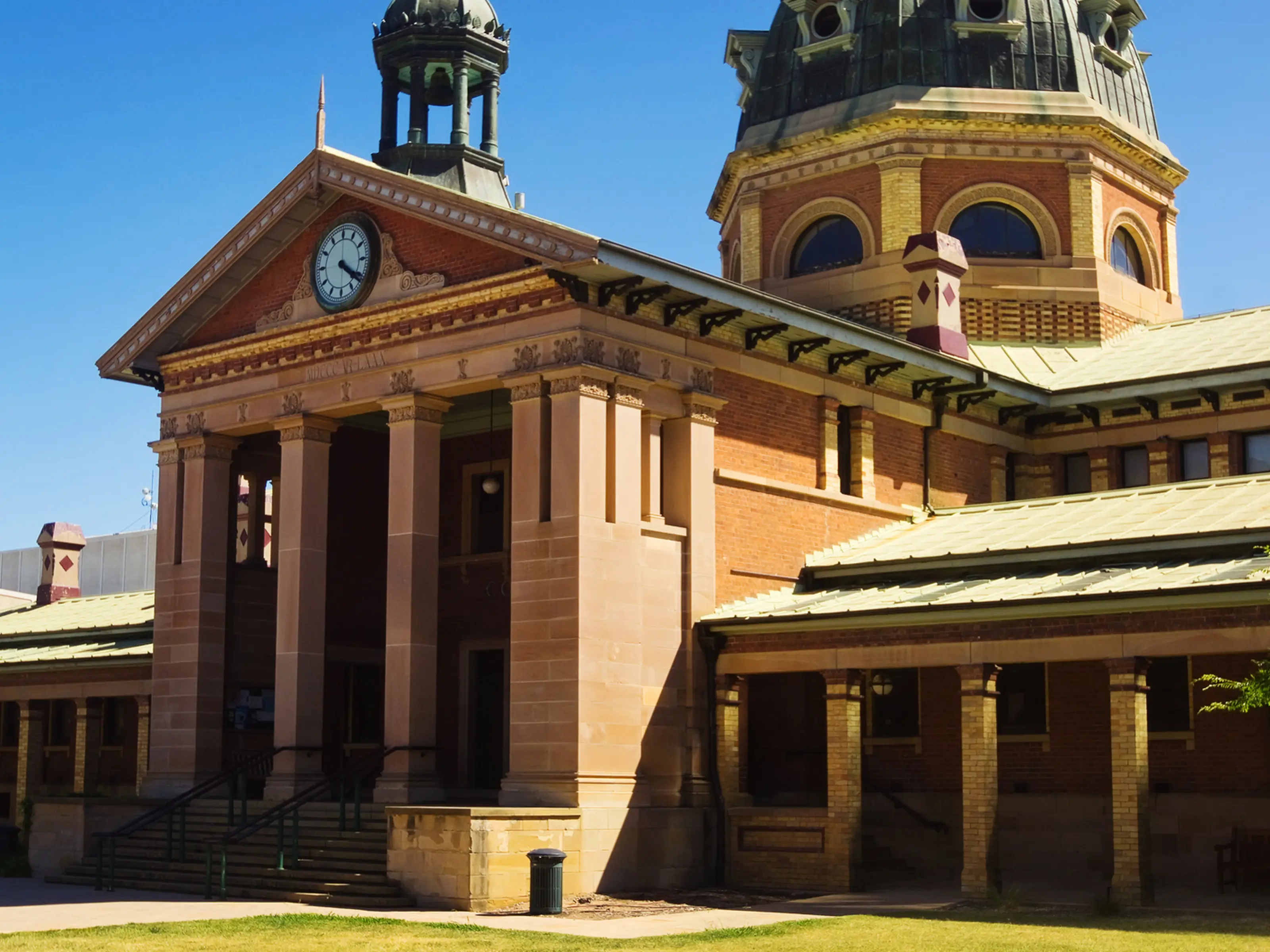 Historic brick building with a clock tower and dome against a clear blue sky, surrounded by a green lawn.
