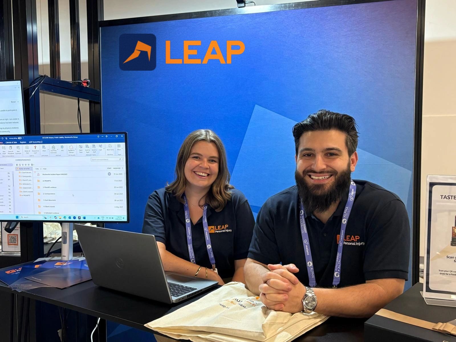 Kate and Aaronw earing LEAP shirts, smilinh at a booth with a laptop and monitor showing software, set against a blue background with the LEAP logo.
