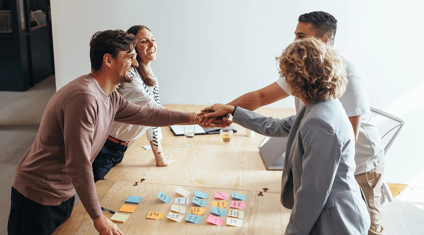 Four people smiling and shaking hands over a table with colorful sticky notes, suggesting a successful meeting or collaboration.