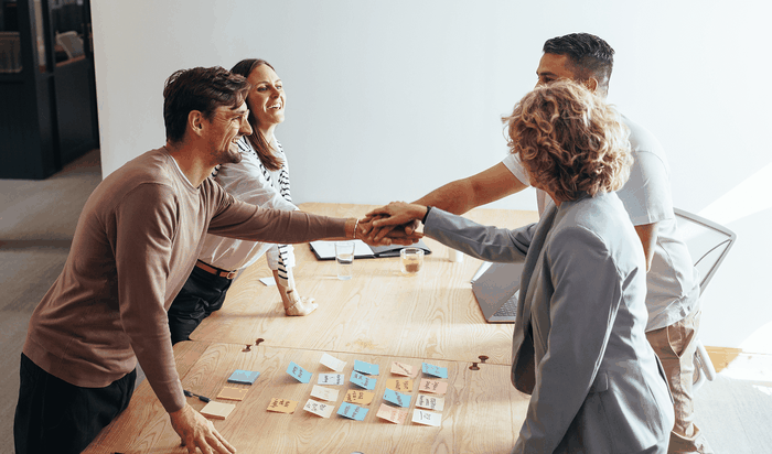 Four people smiling and shaking hands over a table with colorful sticky notes, suggesting a successful meeting or collaboration.