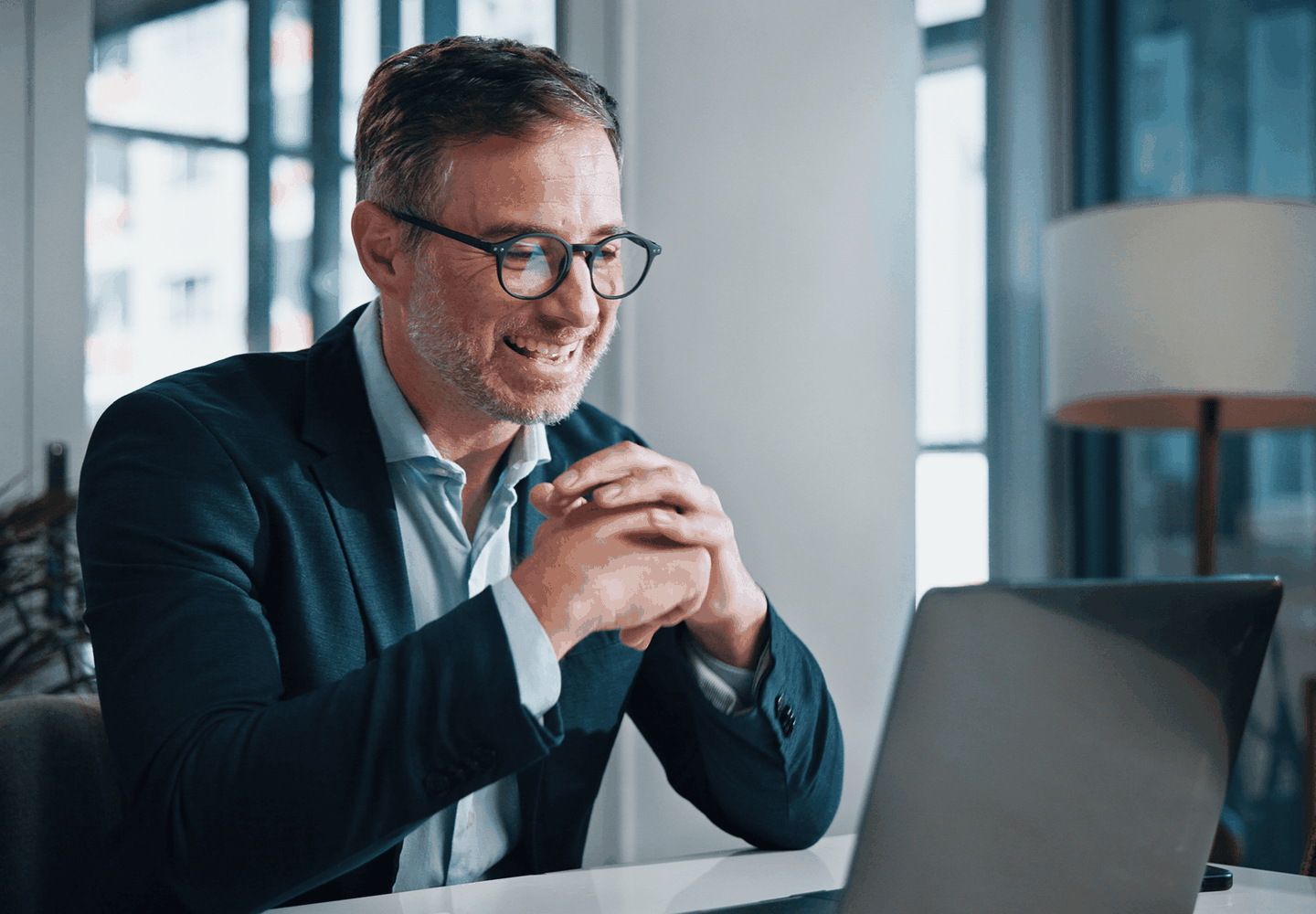 Smiling man with glasses and a suit, sitting at a desk, engaging in a video call on a laptop, in a bright office setting.