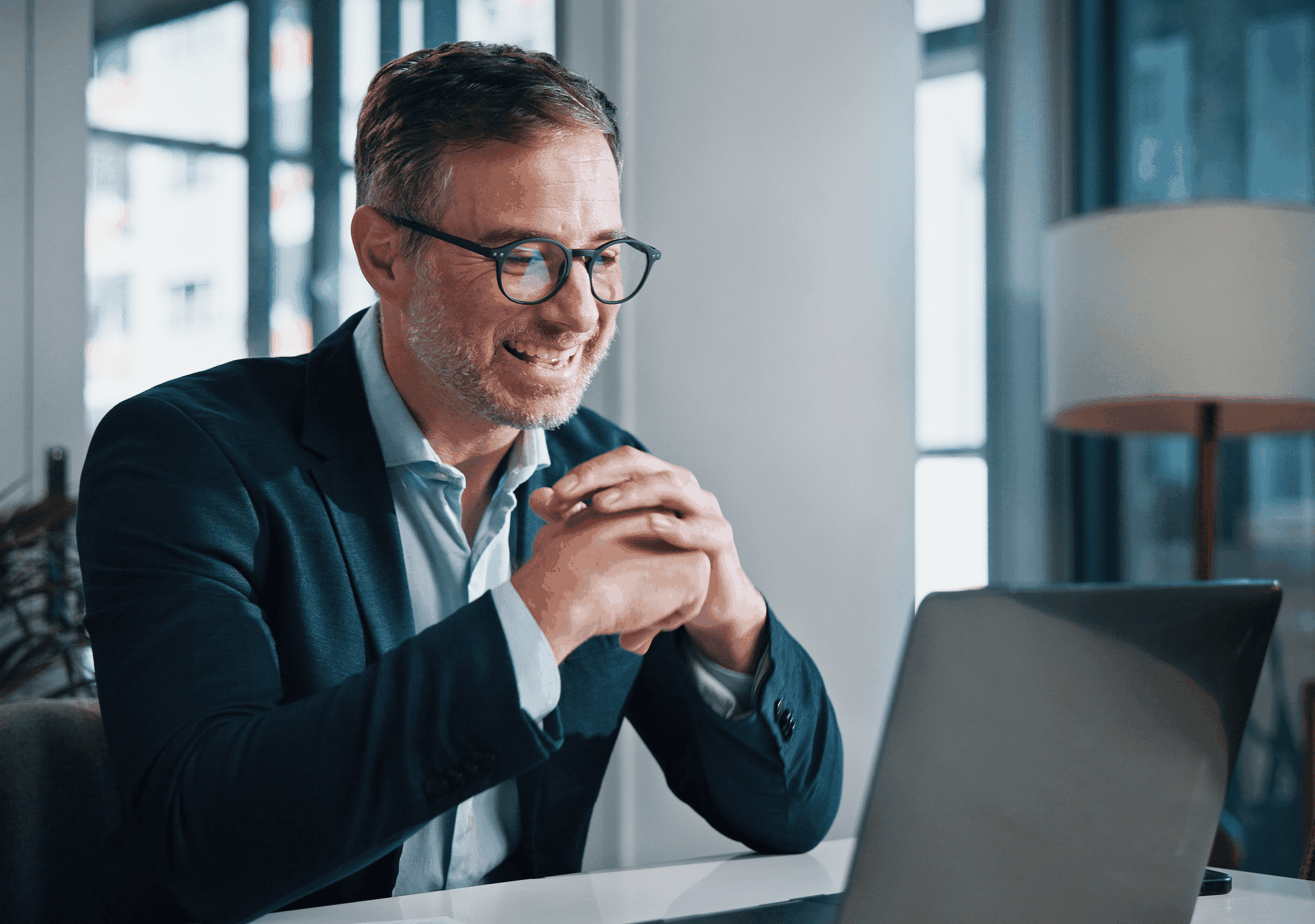 Smiling man with glasses and a suit, sitting at a desk, engaging in a video call on a laptop, in a bright office setting.