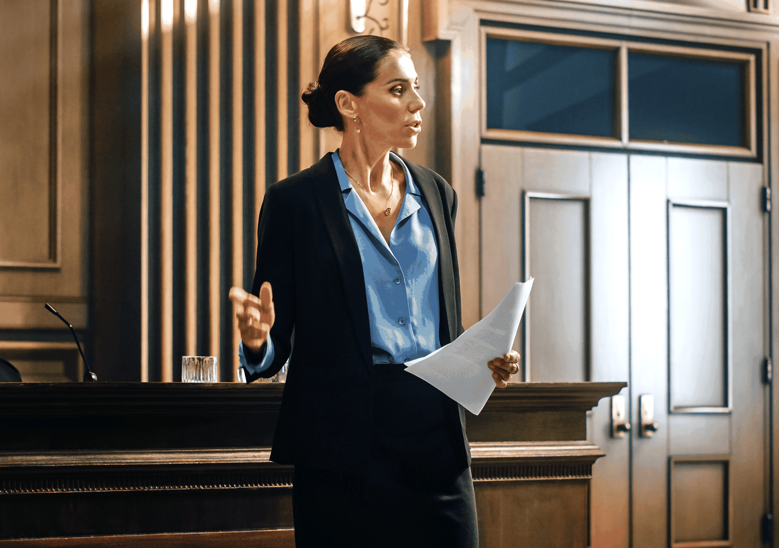 A woman in a courtroom speaks confidently, holding documents, dressed in a blue blouse and black blazer.