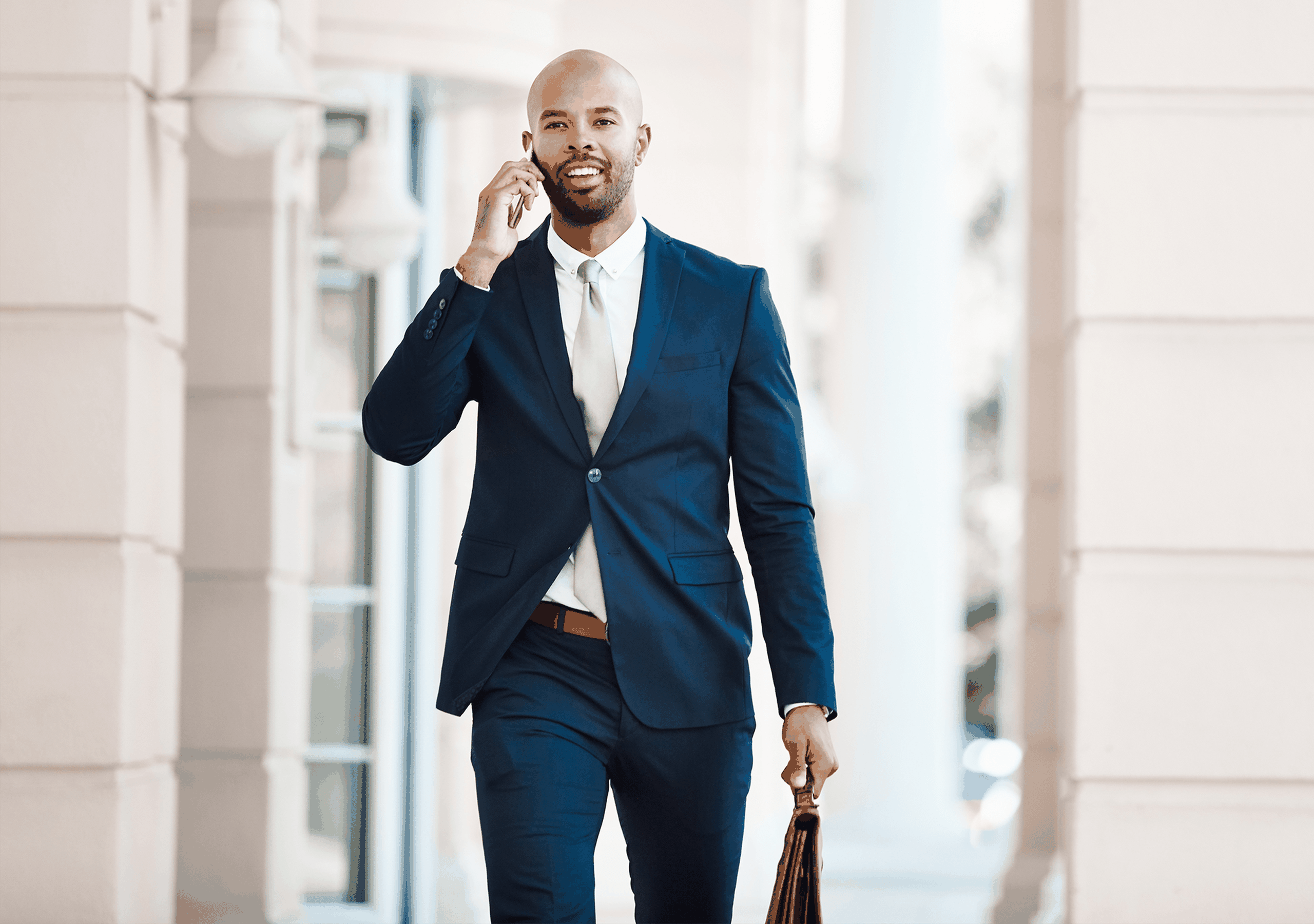 A man in a blue suit walks confidently while talking on his phone and carrying a briefcase in an urban setting.