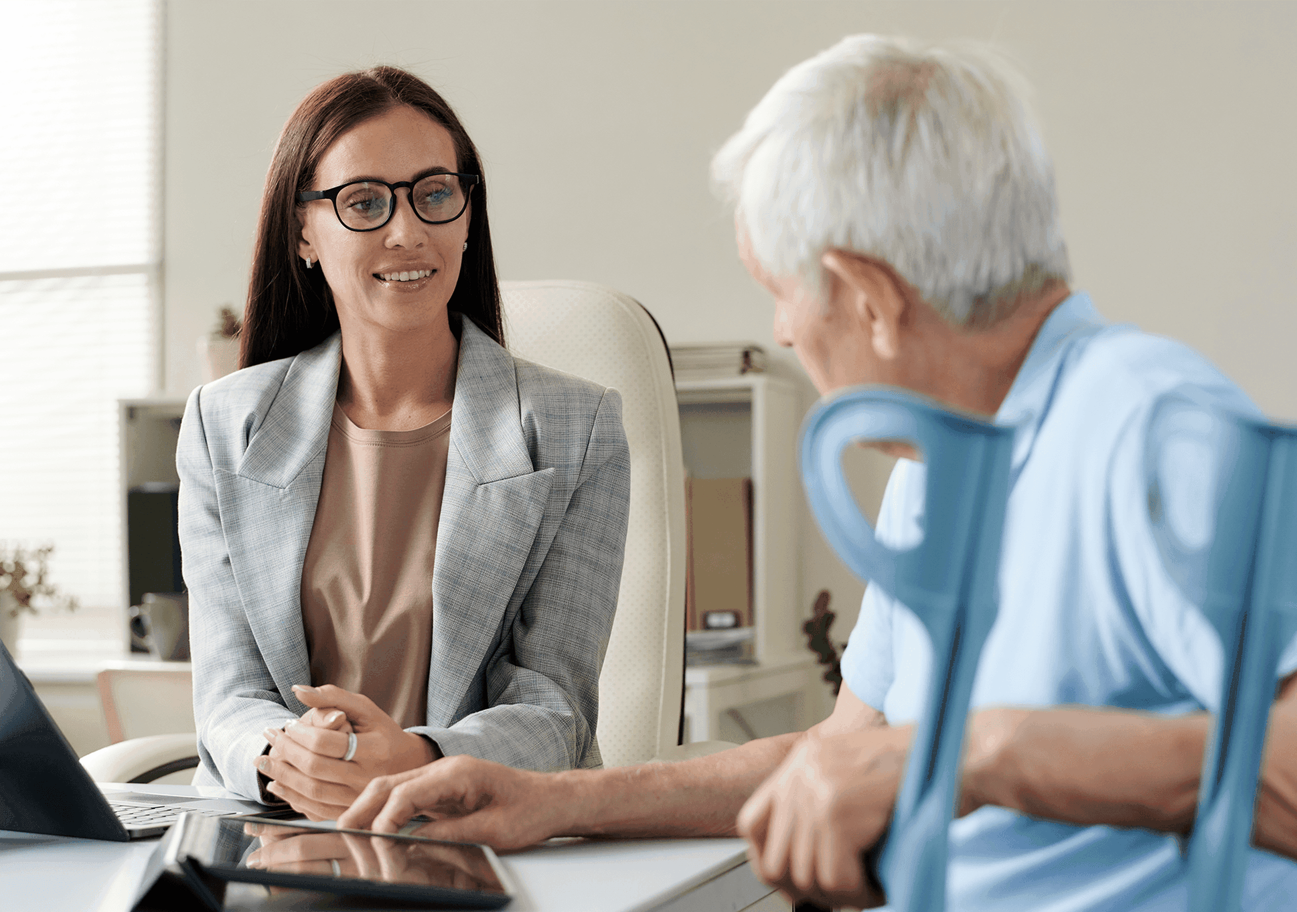 A woman in glasses and a blazer smiles while talking to an older man sitting with crutches in an office setting.