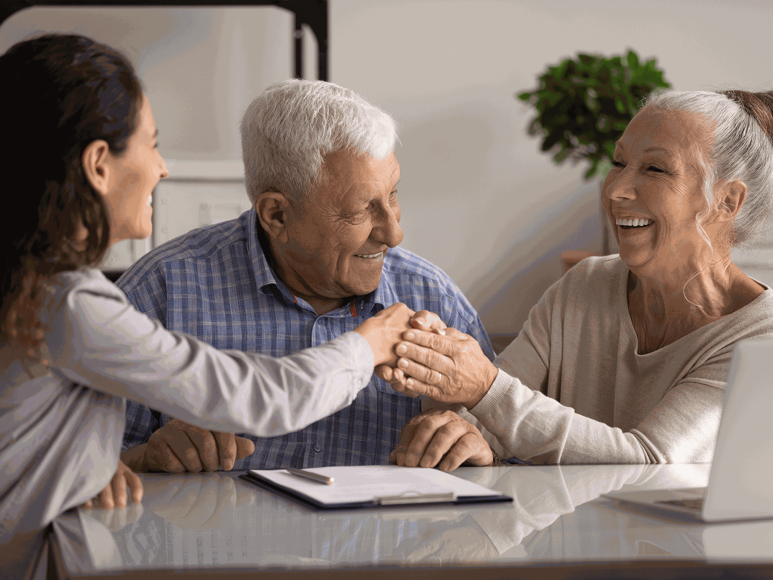 Three people smiling and shaking hands at a table with a notebook, enjoying a friendly conversation. A plant is visible in the background.