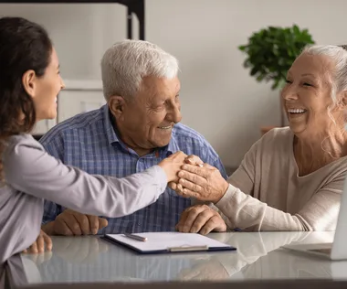 Three people smiling and shaking hands at a table with a notebook, enjoying a friendly conversation. A plant is visible in the background.