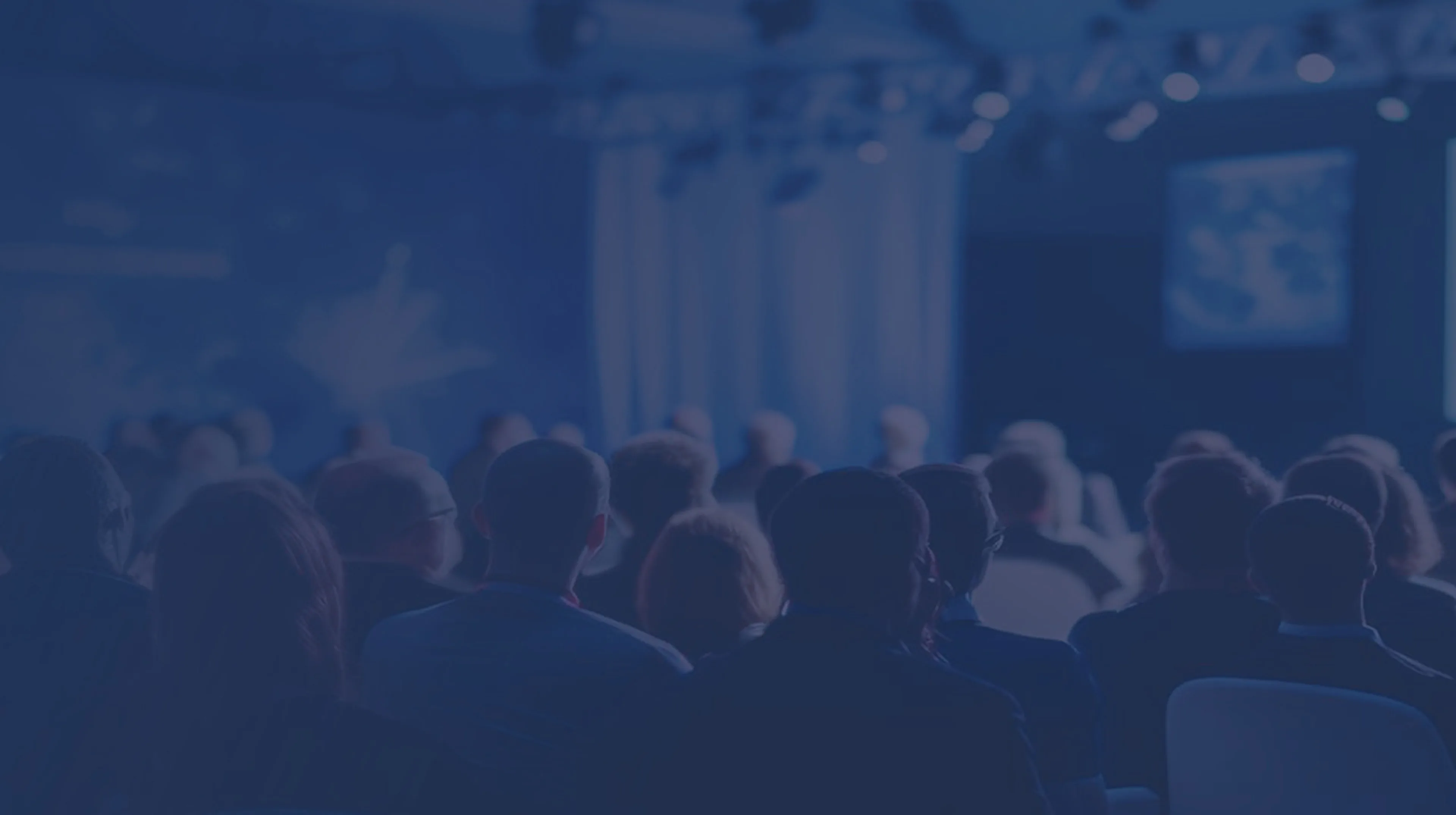 Audience watching a speaker on stage in a dimly lit conference room with a large screen displaying a presentation.