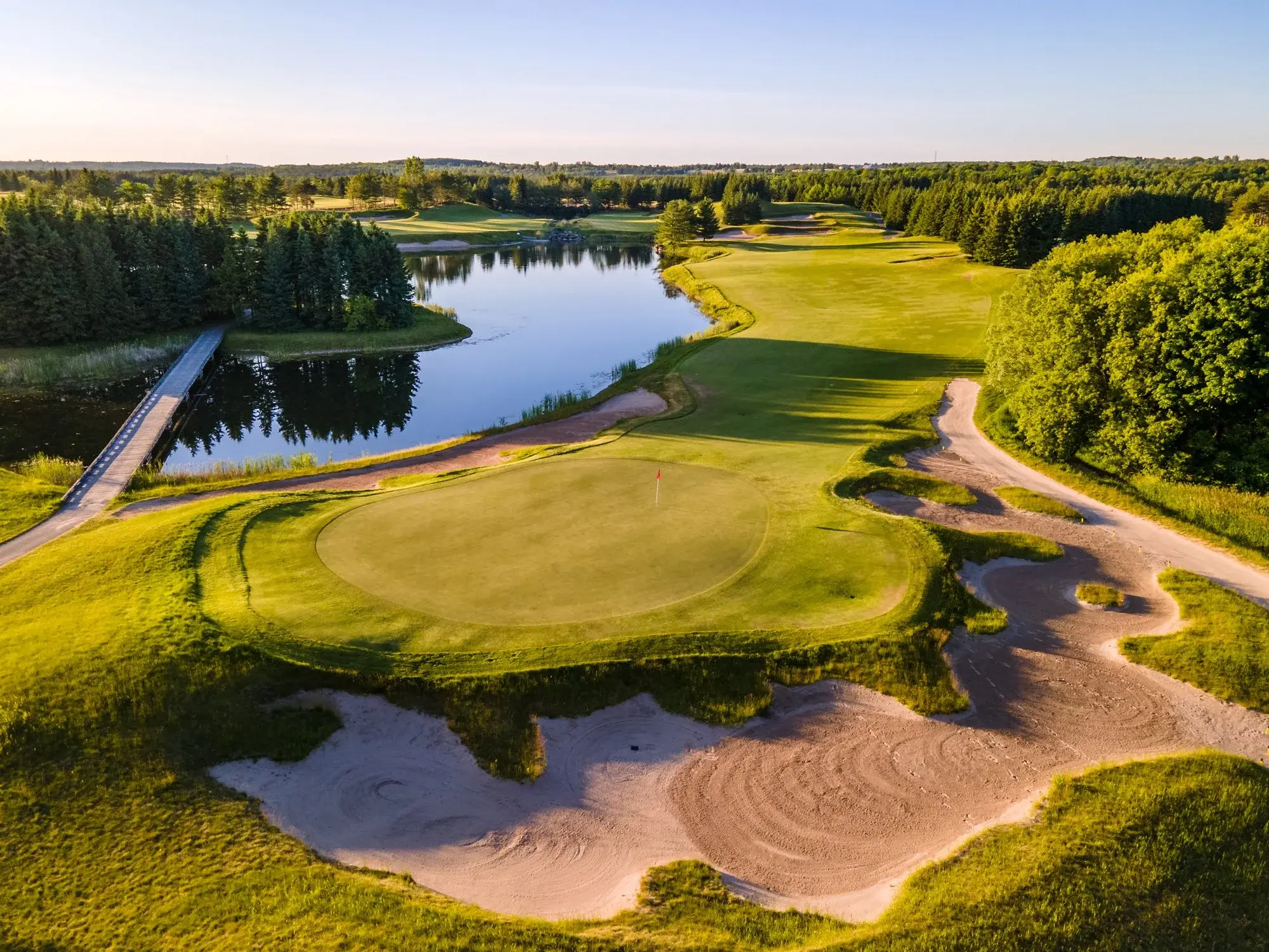 Aerial view of a golf course with a sand trap, lush greenery, a calm lake, and a small bridge under a clear blue sky.