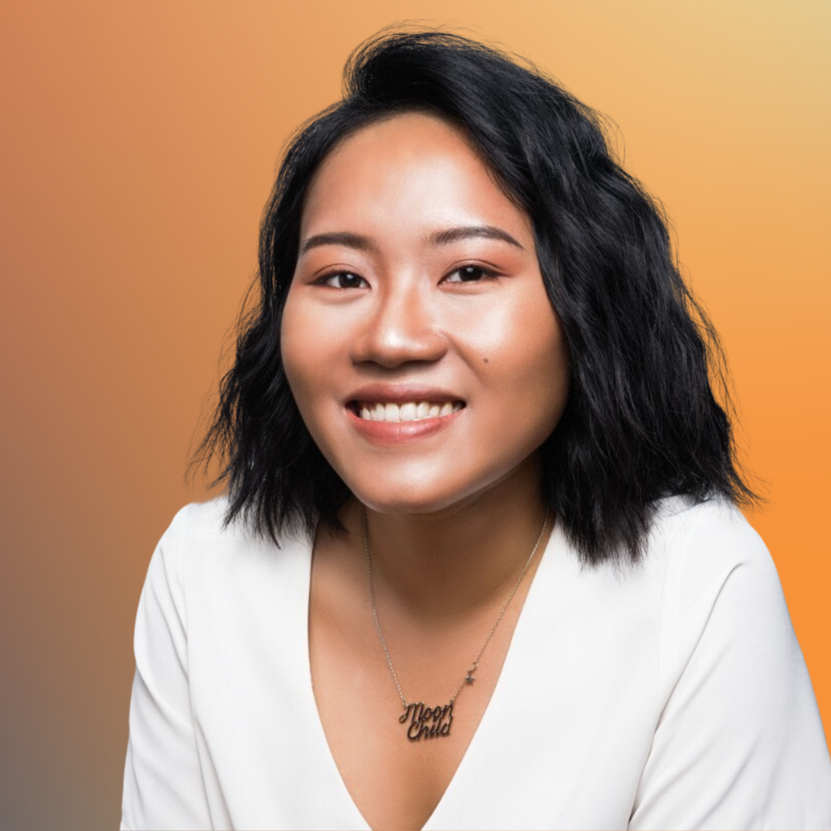 Smiling person with short black hair, wearing a white shirt and "Moon Child" necklace, against a gradient orange background.