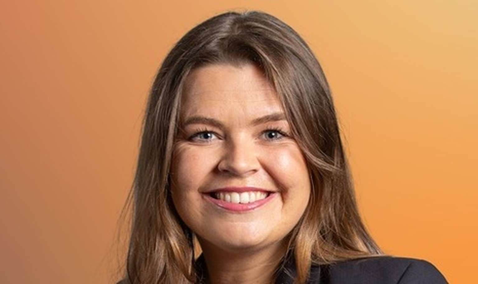Smiling woman with long brown hair, wearing a dark blazer, against a warm orange gradient background.