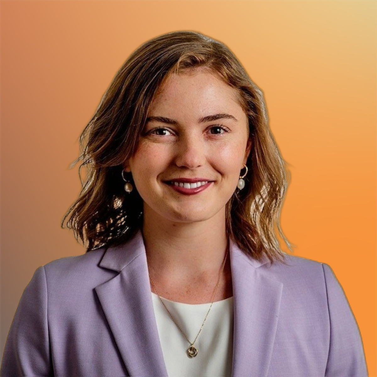 A woman with short wavy hair smiles at the camera, wearing a lavender blazer and white top, against a warm orange gradient background.
